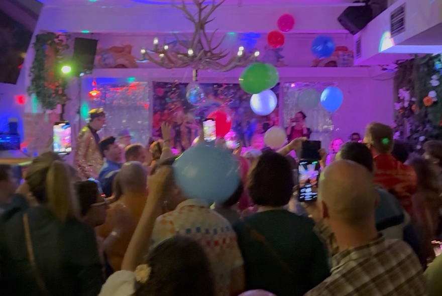 People partying in a colourfully-lit venue with colourful balloons and a band on stage.