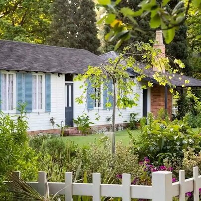 La Trobe Cottage, a white cottage with blue shutters, a dark shingled roof, a brick chimney, and a small porch, surrounded by lush green trees, shrubs, and a white picket fence in the foreground.