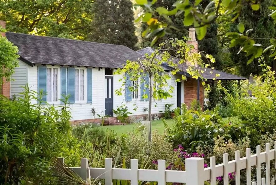La Trobe Cottage, a white cottage with blue shutters, a dark shingled roof, a brick chimney, and a small porch, surrounded by lush green trees, shrubs, and a white picket fence in the foreground.