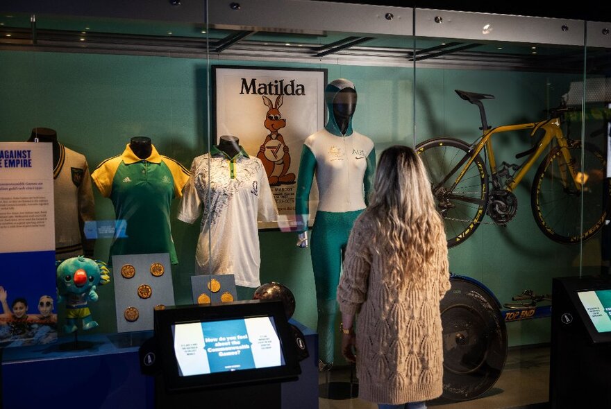 A person viewed from behind viewing a museum display of different sporting outfits and accessories.