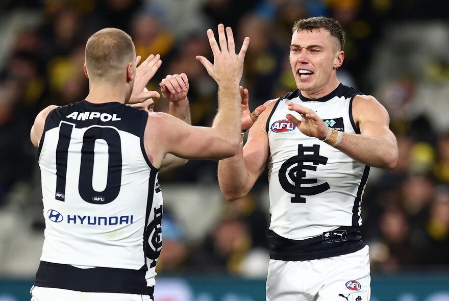 Two Carlton AFL players celebrating on the field after having scored, they face each other and are high-fiving their hands, with blurred fans in the stadium in the background