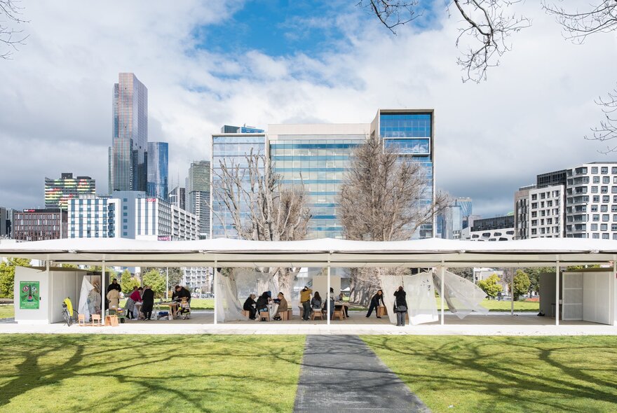 MPavilion, an open-concept building with a flat roof structure surrounded by grass and trees in front of the Melbourne cityscape.
