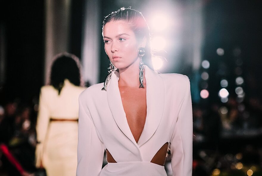 Fashion model walking down a catwalk runway wearing a tailored jacket, spotlit from behind.