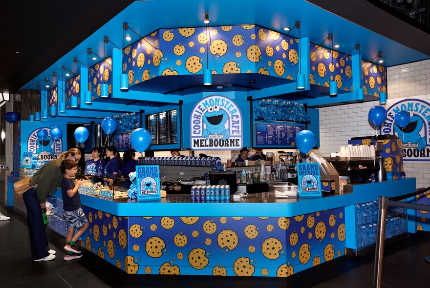 The blue and yellow exterior of the Cookie Monster cafe kiosk at Melbourne Central, with an adult and child looking at the selection of food on display.