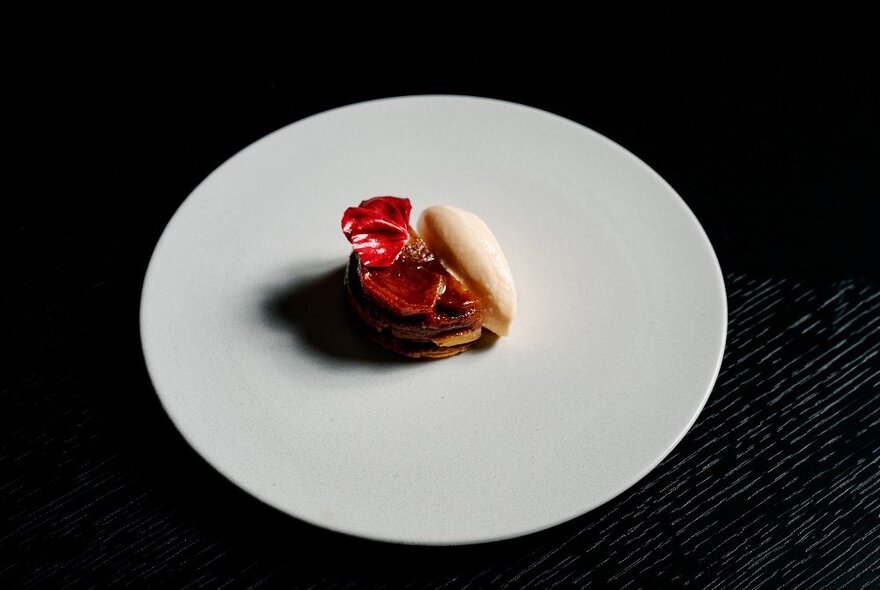 A large white plate with a small round serving of a Japanese delicacy with red petal-like garnishes and a scoop of cream.