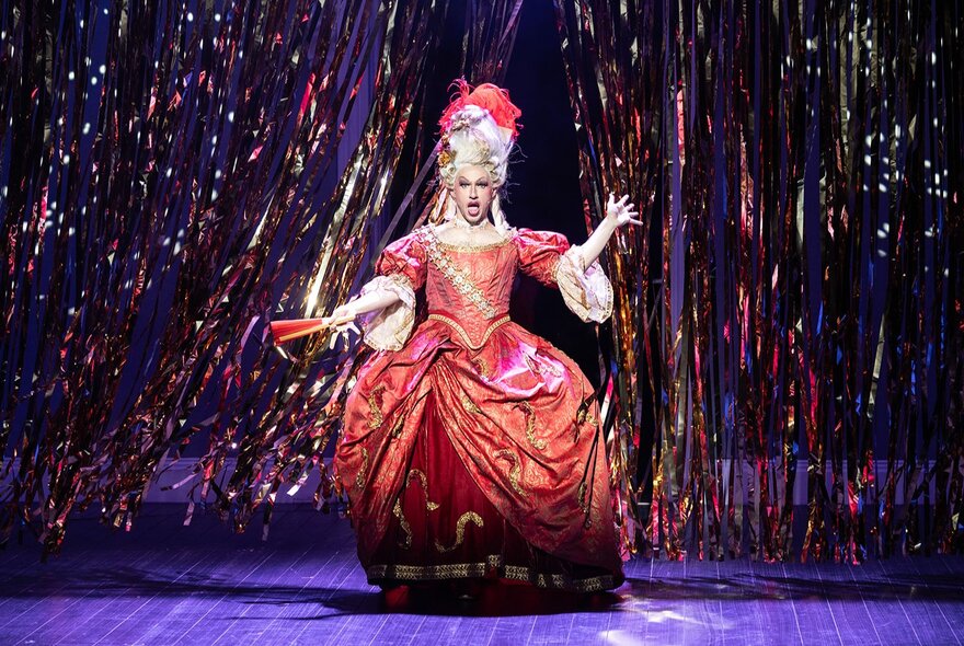 An opera singer in an orange ballgown waling on stage with coloured tinsel in the background. 