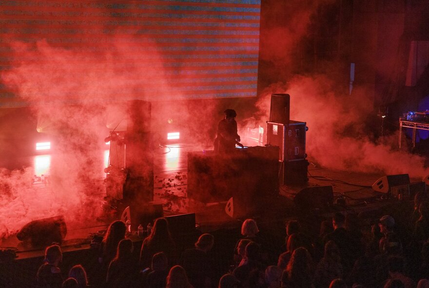 A live musical performance, with red dramatic stage lighting with atmospheric smoke or haze and an audience watching.