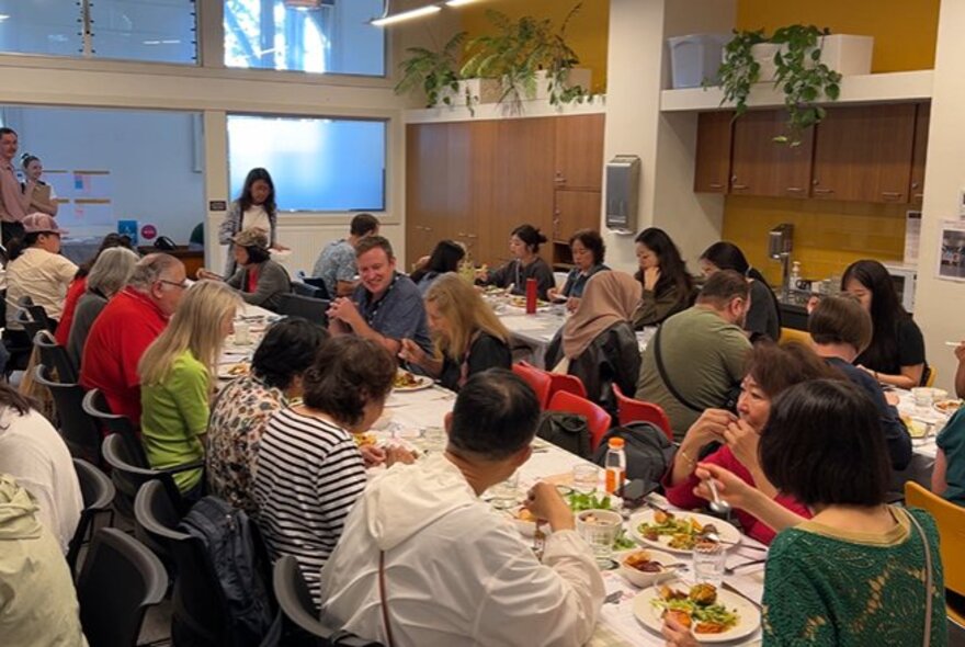 People seated and eating at two long trestle tables inside a community room at the Kathleen Syme library in Carlton.