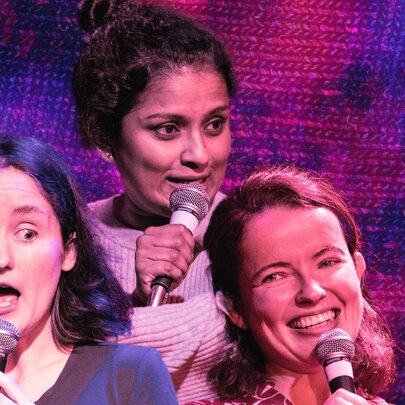 Three female comedians speaking into microphones with varied facial expressions.