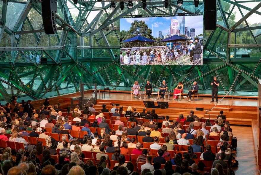 People in rows of seats at The Edge,  listening to a panel of speakers on the stage before them, a large screen in front of the geometric windows.