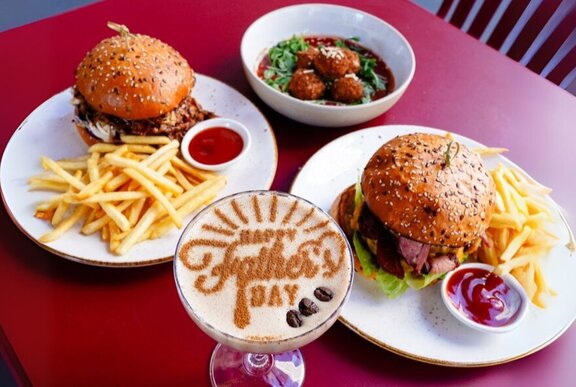 Looking down at a table with two plates of burgers and chips, and a cocktail with the words Father's Day dusted in cocoa on the surface of the drink.