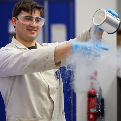 A young man in a white lab coat and safety goggles pours a tin of smoking liquid onto a blue gloved hand in a science laboratory, a fire extinguisher visible on the wall. 