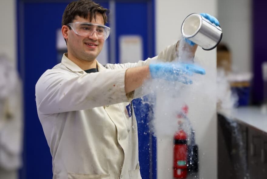 A young man in a white lab coat and safety goggles pours a tin of smoking liquid onto a blue gloved hand in a science laboratory, a fire extinguisher visible on the wall. 