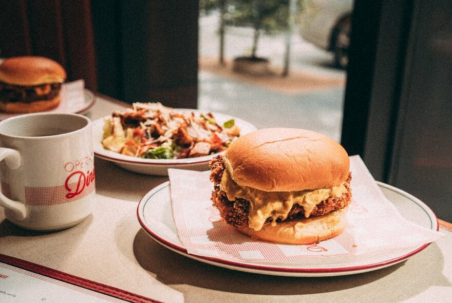 A burger and salad on plates next to a mug, on a cafe table.