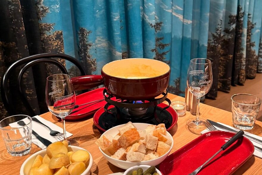 A raclette dish with melted cheese and bowls of bread and potatoes for dipping on a table with wine glasses and red plates.