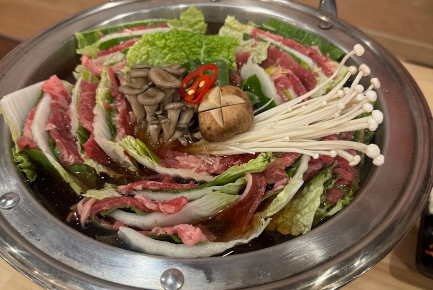 A silver bowl of thinly slice raw meat, vegetables and enoki mushrooms ready to be cooked in a Korean hot pot.