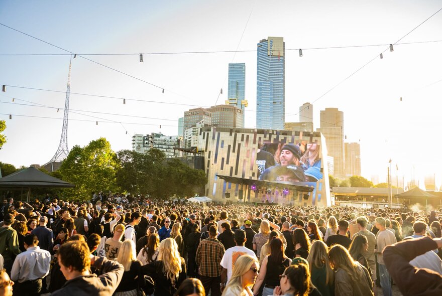 A large crowd of people at Melbourne's Fed Square, watching a film on a large screen; Melbourne's city buildings behind.