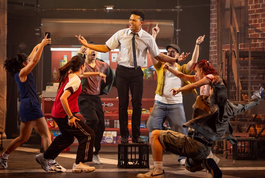 A cast of actors dancing and singing in a musical theatre production on a stage, surrounding a central performer who is standing on an upturned milk crate.
