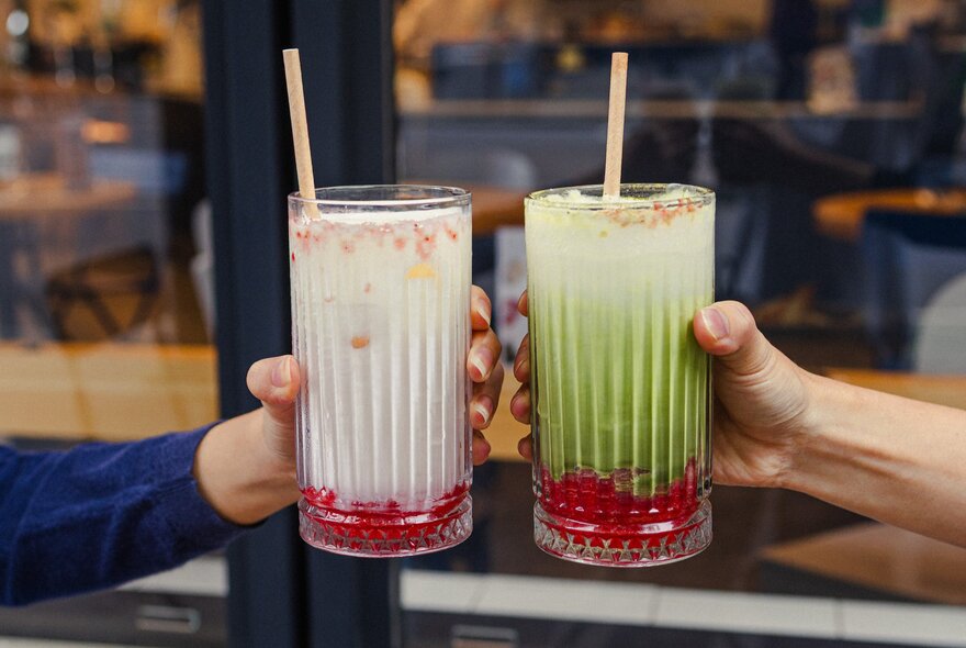 Two hands holding up two long iced drinks with straws in front of a cafe window.