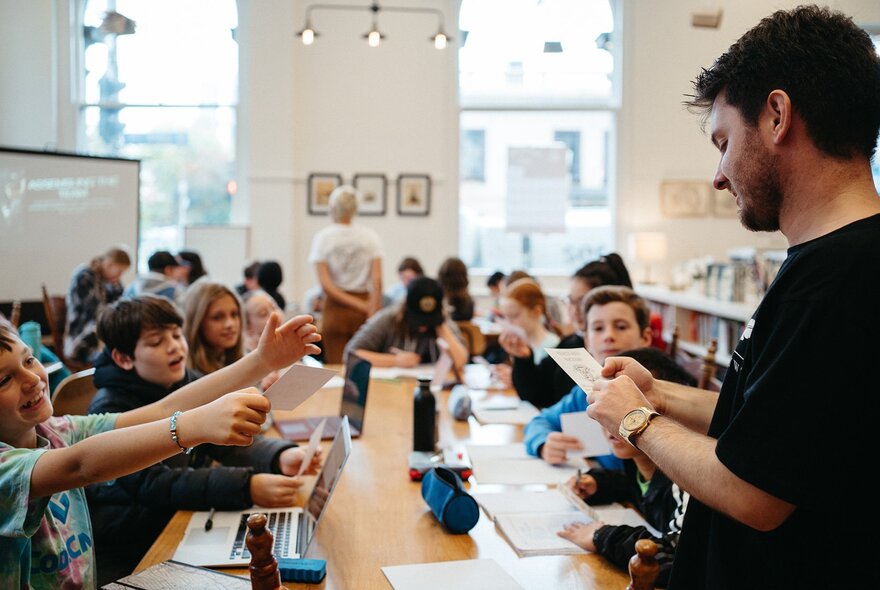 Kids in a classroom setting with a teacher looking on and large windows looking out at buildings and trees.