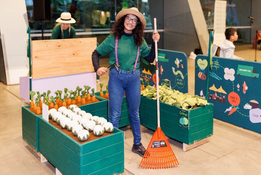 A woman in a typical gardening outfit amongst three mock vegetable garden beds in a gallery, holding a bright orange rake.