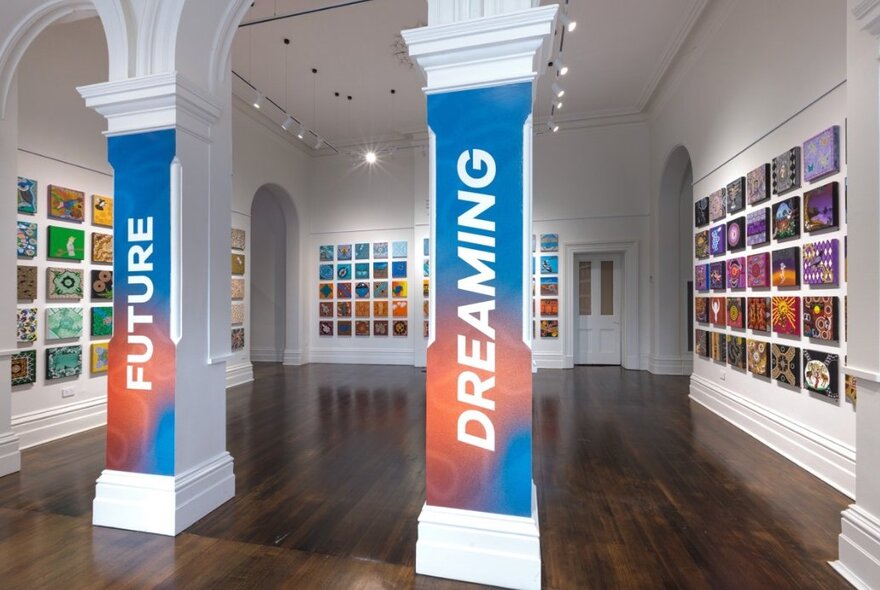 An art gallery room with the entrance pillars wrapped in colourful banners reading 'FUTURE' and 'DREAMING', with grid patterns of small paintings covering the walls.