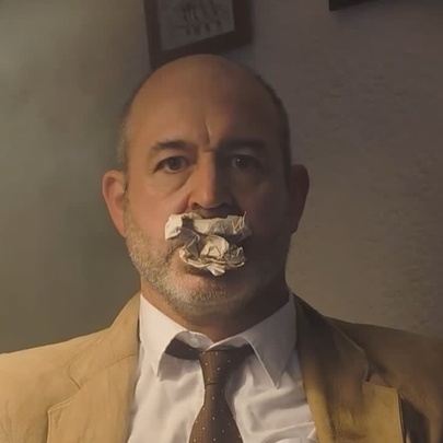 A man in a beige jacket and brown tie, staring at the camera with a scrunched up ball of paper in his mouth.