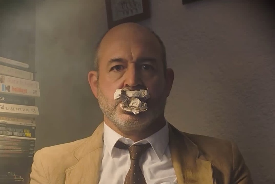 A man in a beige jacket and brown tie, staring at the camera with a scrunched up ball of paper in his mouth.