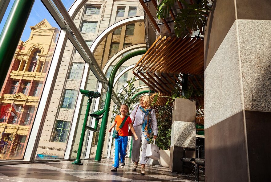 Adult and child walking through glass corridor linking buildings with sunshine streaming in.