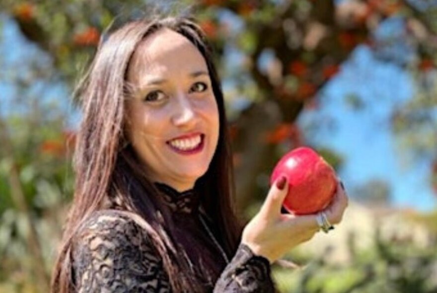 Smiling woman with long dark hair holding a red apple in front of a tree.