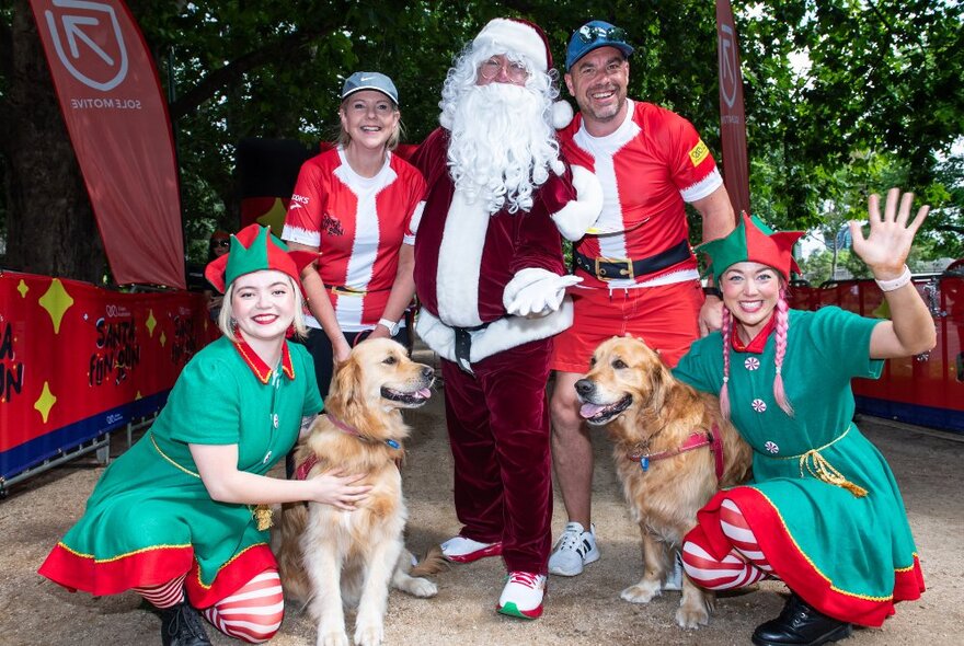 Two runners in Santa red t-shirts, posing for a photograph with Santa and two Elves and two dogs, outside in a park.