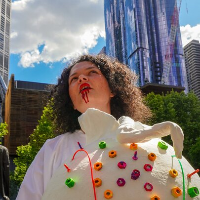 An individual with curly hair wearing a white, textured costume adorned with colourful round elements and fake blood dripping from their mouth, standing outdoors in front of a city skyline.