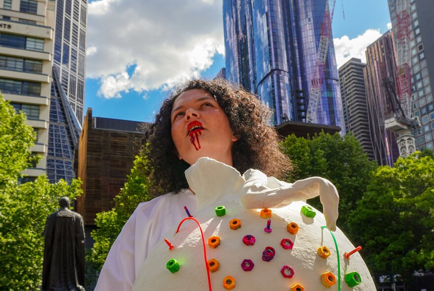 An individual with curly hair wearing a white, textured costume adorned with colourful round elements and fake blood dripping from their mouth, standing outdoors in front of a city skyline.