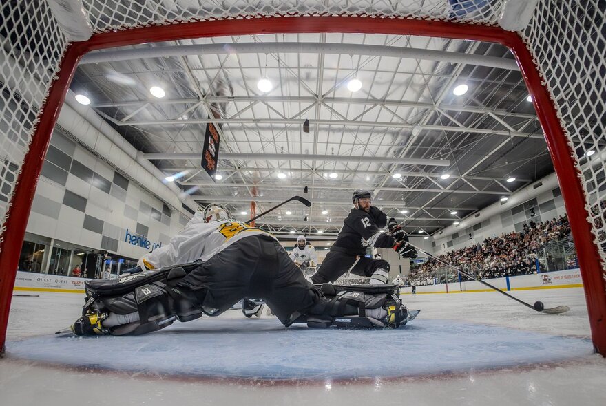 View of ice hockey players and the goal area of an ice hockey rink from ground level and behind the goal nets.