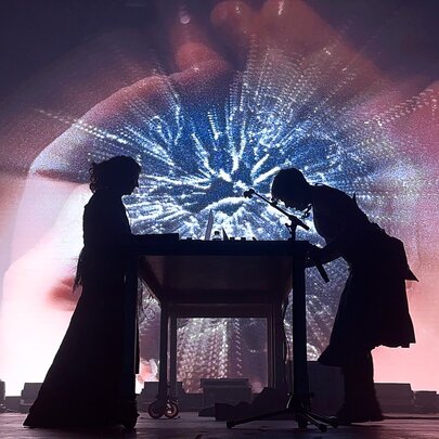 An electronic music performance duo in silhouette performing live against a luminous  backdrop.