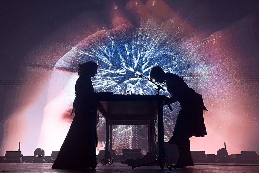 An electronic music performance duo in silhouette performing live against a luminous  backdrop.