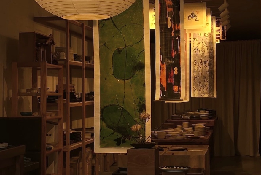 An interior view of a shop and gallery, featuring hanging textile panels or wall scrolls in a minimalist, warm-toned setting, a large bookshelf against one wall.