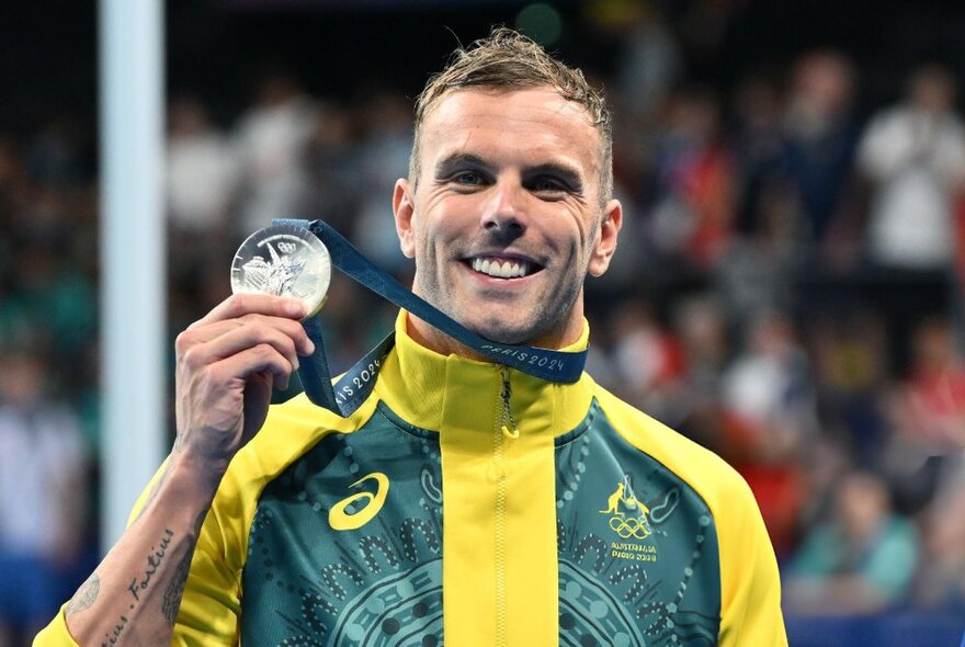 Olympic swimmer, Kyle Chalmers, in Australian colours holding up an Olympic Medal with a blurred crowd in the background.