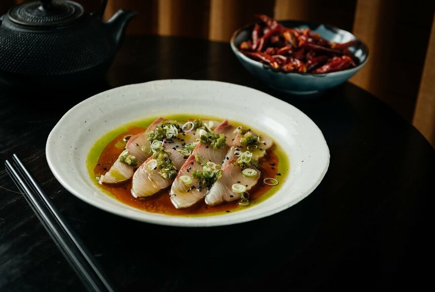 A plate of sashimi with a bowl of chillies in the background. 