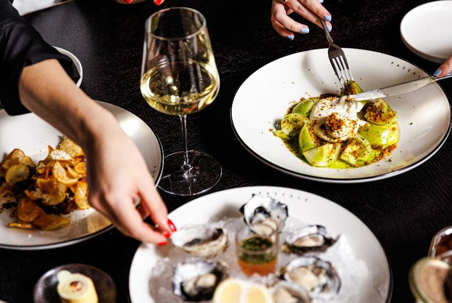 People's hands and plates of pasta and oysters on a table with glasses.