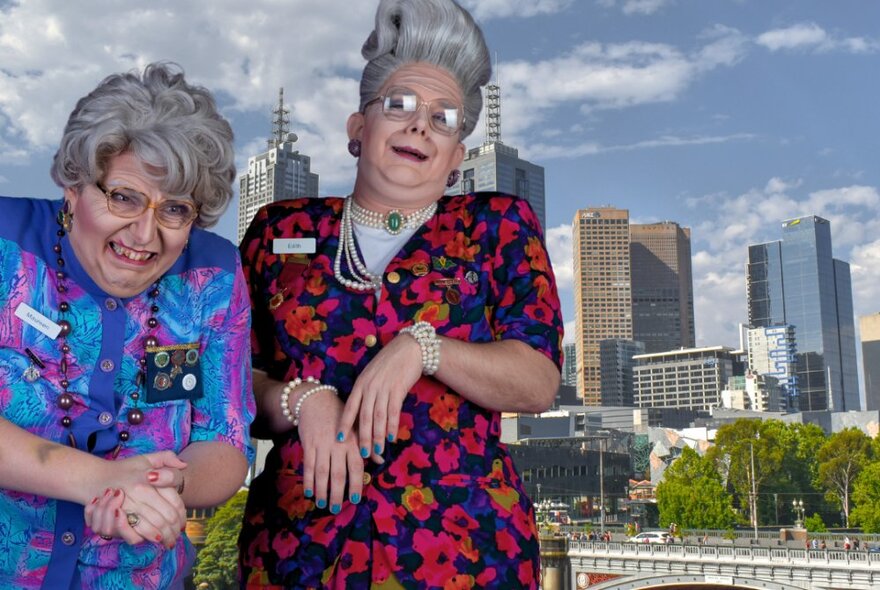 Two men dressed as elderly ladies wearing wigs, dresses and jewels with the city skyline in the background.