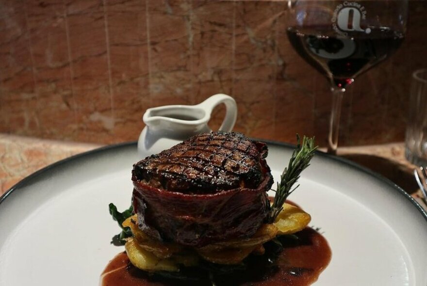 Meat dish on a white plate with a gravy jug and glass of red wine.