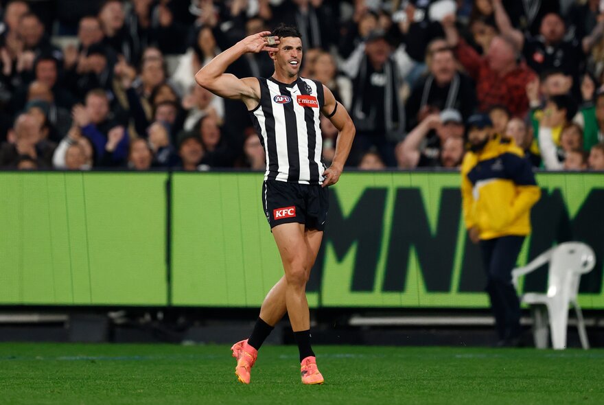 A Collingwood AFL football player on the field during a match, celebrating a goal, with cheering fans in the stadium behind him.