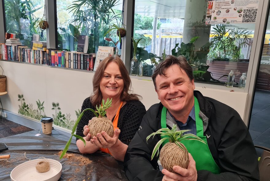Two class participants in coloured aprons holding up kokedama balls and smiling at a craft table. 