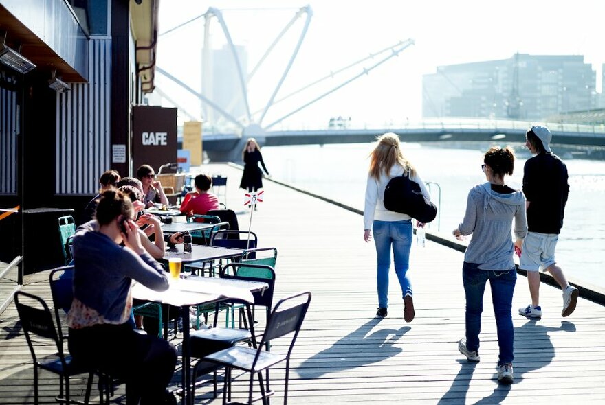 People seated at outdoor cafe tables by a river as pedestrians walk past.