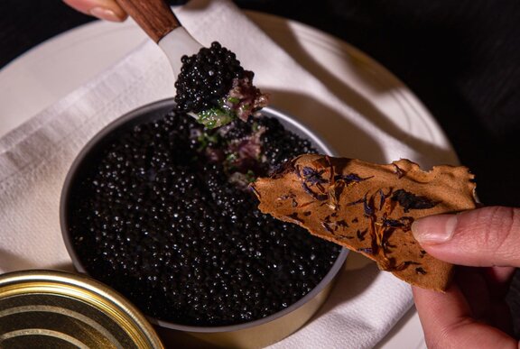A hand dipping a cracker into a tin of black caviar.