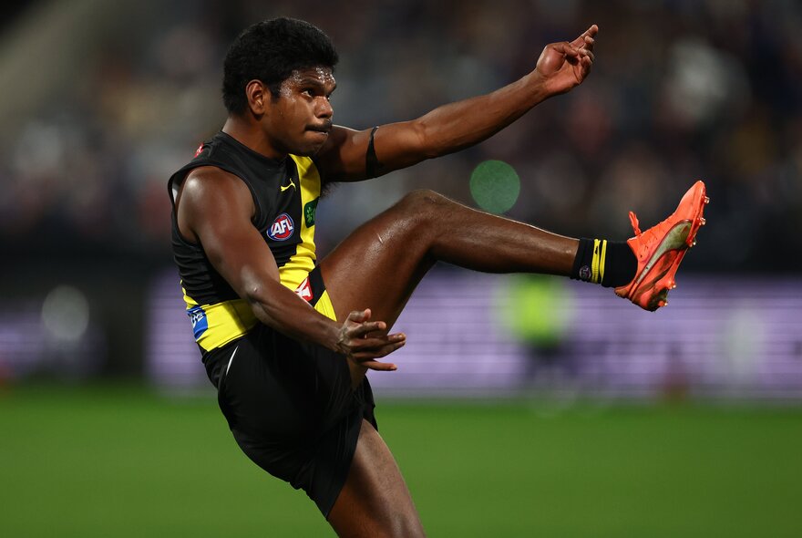 A Richmond AFL football player on the field during a match kicking a ball that is out of sight.