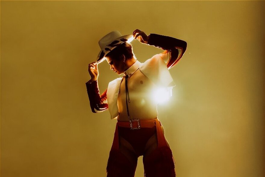 A man with a bare chest wearing a white vest, chaps and a cowboy hat, spot lit from behind against a mustard coloured background.