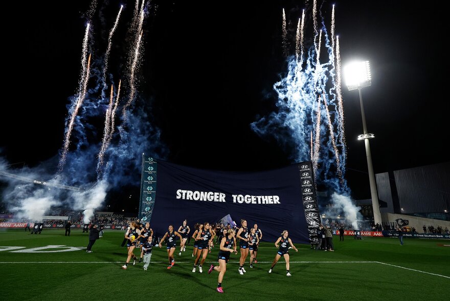 Carlton AFLW players running through a banner reading stronger together while fireworks go off at night on a footy oval.