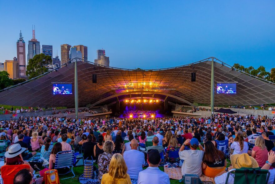 Crowds sitting on the lawn watching a performance at the Sidney Myer Music Bowl where a large roof covers the stage, with the city behind.